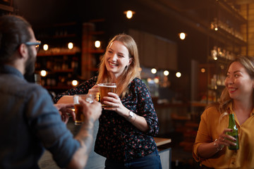 Sticker - Smiling beautiful blonde leaning on bar counter, holding pint of beer and talking to her friend. Nightlife.