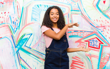 Wall Mural - young pretty african american woman smiling, feeling happy, positive and satisfied, holding or showing object or concept on copy space against graffiti wall