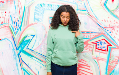 Wall Mural - young pretty african american woman smiling cheerfully and casually, looking downwards and pointing to chest against graffiti wall