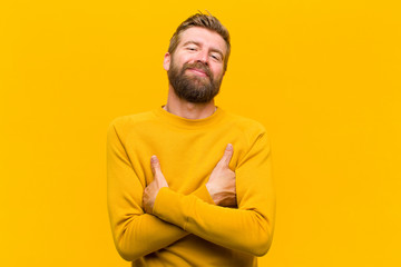 young blonde man laughing happily with arms crossed, with a relaxed, positive and satisfied pose against orange wall