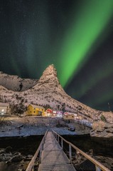 Sticker - Wooden pier going towards a village on a snow covered hill under the beautiful auroras in the sky