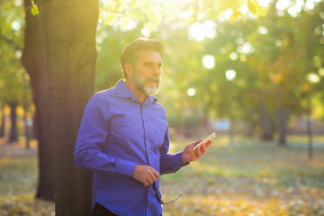 Wall Mural - Happy smiling businessman with gray beard wearing blue shirt and using modern smartphone near tree in the park morning, successful employer to make a deal while standing near tree 