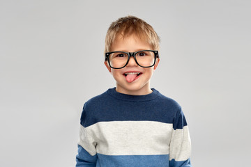 Wall Mural - school, education and vision concept - portrait of smiling little boy with crookedly worn glasses showing tongue over grey background