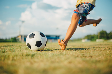 A boy kicking a ball with bare foot while playing street soccer football on the green grass field for exercise in community rural area under the twilight sunset.