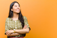 Young Pretty Hispanic Woman Feeling Happy, Proud And Hopeful, Wondering Or Thinking, Looking Up To Copy Space With Crossed Arms Against Brown Wall