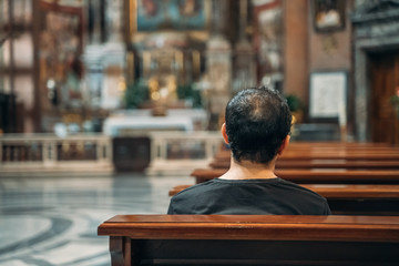 Believer sits and prays on bench inside old big temple or church, view from back, selective focus