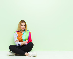 Wall Mural - young blonde woman smiling to camera with crossed arms and a happy, confident, satisfied expression, lateral view sitting on the floor