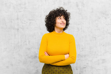 young pretty afro woman feeling happy, proud and hopeful, wondering or thinking, looking up to copy space with crossed arms