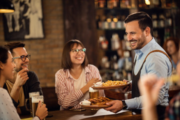 Happy waiter serving food to guest who are drinking beer in a pub.