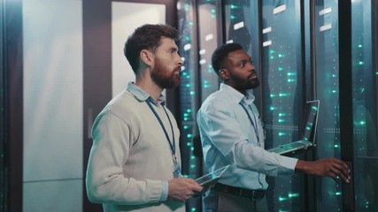 In modern data center two server technicians working in server rack room inspecting software network using notebook and digital tablet.