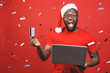 © denis_vermenko - Portrait of an excited young african american man dressed in santa claus costume standing isolated over red background, holding laptop computer, showing plastic credit card.