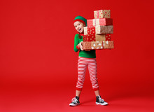 Cheerful Funny Baby In Christmas Elf Costume With Gifts On   Red Background.