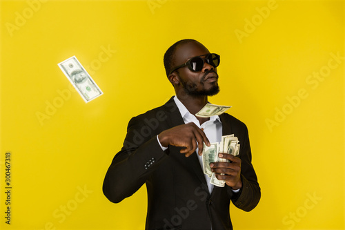 Fotografía bearded solid young afroamerican guy is throwing out dollars from one hand, wear