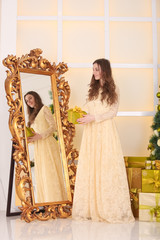 beautiful slim caucasian adult woman in long evening yellow dress holding gift box with bow and looking at the mirror while celebrating christmas in her white room