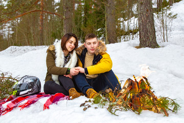  A man and a woman sitting by a fire in the winter forest. Lovers hold hands and look at the fire. Winter vacation together. Romantic journey through the forest. Winter rest in nature.