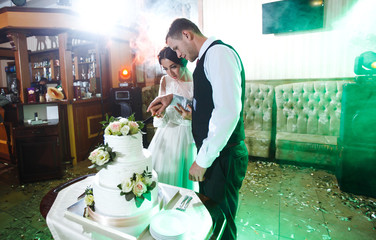 Wall Mural - Elegant and stylish wedding cake with flowers. Newlyweds standing near a large wedding cake. Bride and a groom is cutting their wedding cake on wedding banquet in restaurant. Marriage. Together.