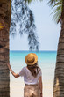 © allenkayaa - A young woman under a palm tree on the shore of the Gulf of Thailand. Woman in a hat looks at the sea