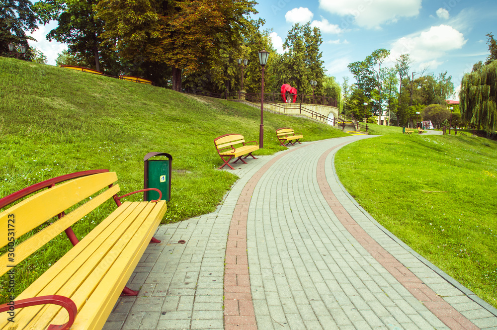 Beautiful park with an alley and benches and a green meadow.  Blue sky with clouds, grass and trees - obrazy, fototapety, plakaty 