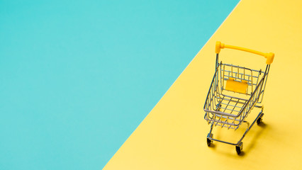 Empty miniature shopping cart on blue and yellow background. Toy trolley on bright colorful background, copy space for text or design.