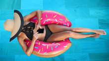 Summer Vacation Woman In Bikini On Inflatable Donut Mattress Using Mobile Cell Phone In Swimming Pool. Top View Of Girl Relaxing Sunbathing Enjoying Travel Holidays At Resort Pool. Luxury Lifestyle.