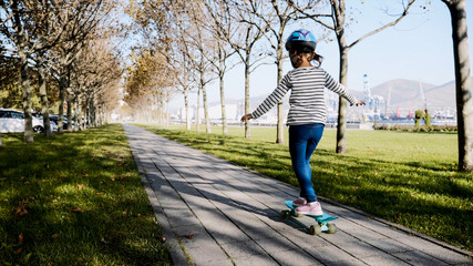 Little girl is slowly skateboarding in protective helmet in the park on path at sunny autumn weather.