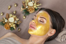 Beautician Makes A Golden Mask To Rejuvenate The Skin To Beautiful Woman. Next To Her Are Christmas Decorations.New Year's And Cosmetology Concept.