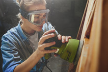 Young Woman In Safety Glasses Polishes A Wooden Bed With Random Orbit Sander, DIY Concept