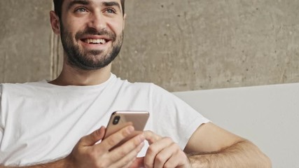 Wall Mural - Close up view of handsome happy young brunet man smiling and using smartphone while sitting on couch at home