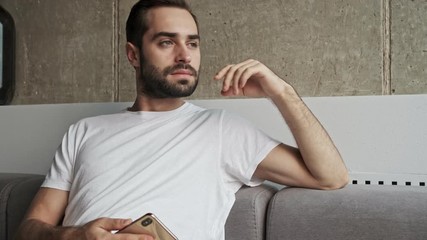 Wall Mural - Handsome pensive young brunet man with smartphone feeling sad and looking at the window while sitting on couch at home