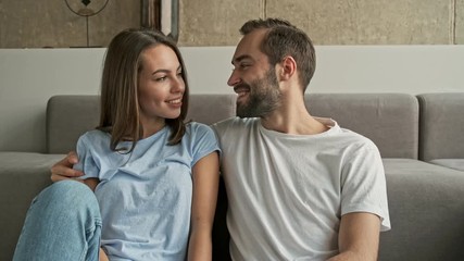 Wall Mural - Handsome happy young lovely couple hugging and talking while looking at each other sitting on the floor near the couch at home