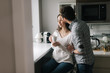 © dikushin - Confident young handsome man kisses his girlfriend in th kitchen by the window. Daily life of a male and female in apartment.