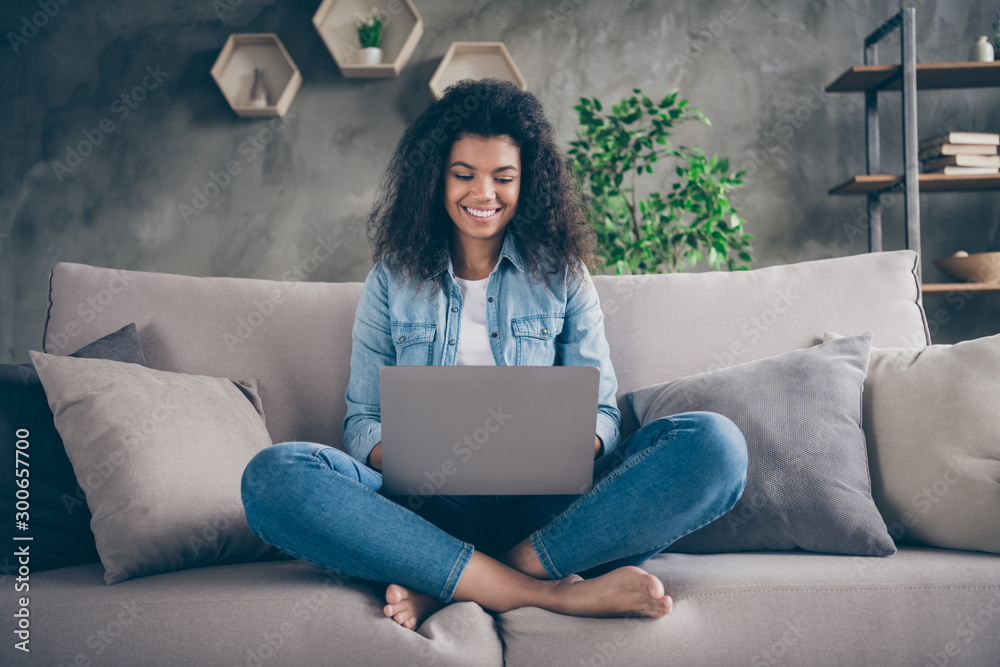 Photo of pretty amazed dark skin curly lady notebook on knees crossed legs texting friends read good news sitting comfy couch casual denim outfit living room indoors - obrazy, fototapety, plakaty 