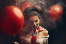 Creepy Beautiful Woman In The Image Of A Scary Clown. Carnival Outfit White Dress With Red Buboes. Close-up Portrait. Bloody Smile, Obsessed With Crazy Eyes. Creative Art Makeup. Halloween Celebration