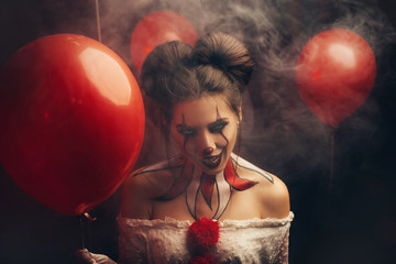 Creepy beautiful woman in the image of a scary clown. Carnival outfit white dress with red buboes. Close-up portrait. Bloody smile, obsessed with crazy eyes. Creative art makeup. Halloween celebration