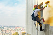 © predragmilos - Industrial rope access worker hanging from the building while painting the exterior facade wall. Industrial alpinism concept image. Top view