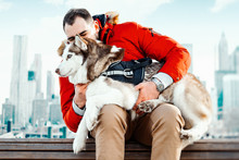 Man And Cute Siberian Husky Dog Sitting On Bench On Cityscape Background. Human And Animal Pure Love. Best Friend.