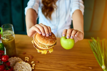 Wall Mural - Beautiful young woman decides eating hamburger or apple in kitchen. Cheap junk food vs healthy diet