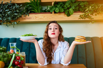Wall Mural - Beautiful young woman decides eating hamburger or fresh salad in kitchen. Cheap junk food vs healthy diet