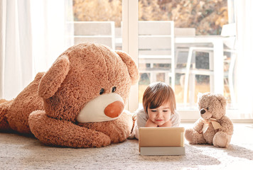 Cute little child watching cartoons on digital tablet device lying on floor with two soft teddy bear toys at home. Modern childhood.