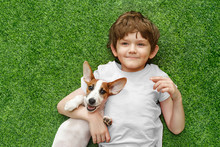 Child Embracing Puppy Jack Russell And Lying On Green Carpet.
