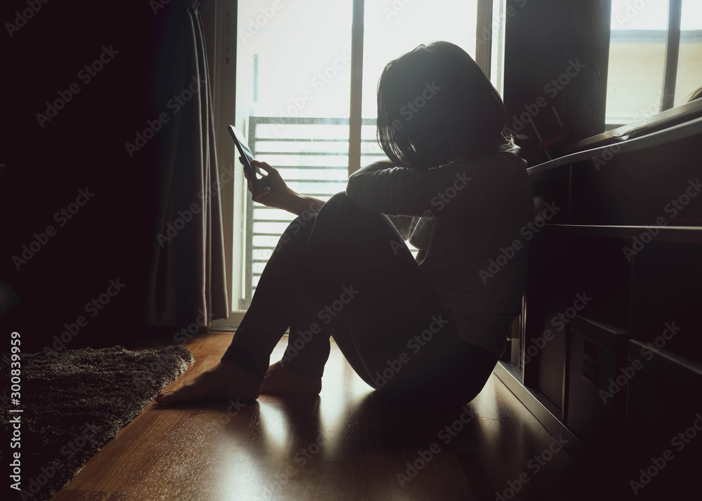 Asian woman sitting on wood floor beside window light in a dark room. Hold and bend head to look at mobile phone screen to check message, chat bad news or wait for a reply by feeling sad or hopeless - obrazy, fototapety, plakaty 
