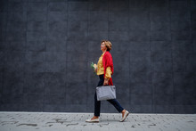 Side View Of Charming Caucasian Fashionable Senior Woman Carrying Bag And Groceries While Passing By Gray Wall.