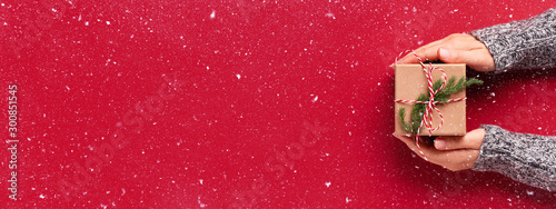 Cuadros en Lienzo Female's hands in pullover holding Christmas gift box decorated with evergreen branch on red background with snow
