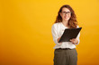 Beautiful young girl in glasses with a clipboard smiles and looks in camera standing isolated on yellow background.