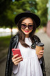 © dianagrytsku - Young asian woman with hat and sunglasses reading using phone in the street. Female asian woman reading news or texting sms on smartphone while drinking coffee on break from work.