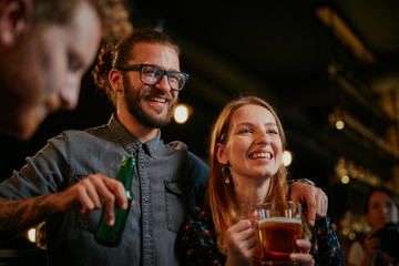 Poster - Handsome caucasian man with eyeglasses hugging his girlfriend and talking to her while standing in a pub and drinking beer. Nightlife.