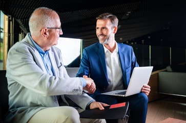 younger and older businessman shaking hands in modern business lounge