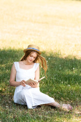 Wall Mural - full length view of beautiful girl in white dress and straw hat reading book while sitting on meadow