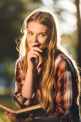 Wall Mural - portrait shot of beautiful girl in casual clothes sitting on meadow in sunlight, holding book and looking away