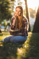 Wall Mural - full length view of beautiful girl in casual clothes sitting on meadow in sunlight, holding book and looking at camera